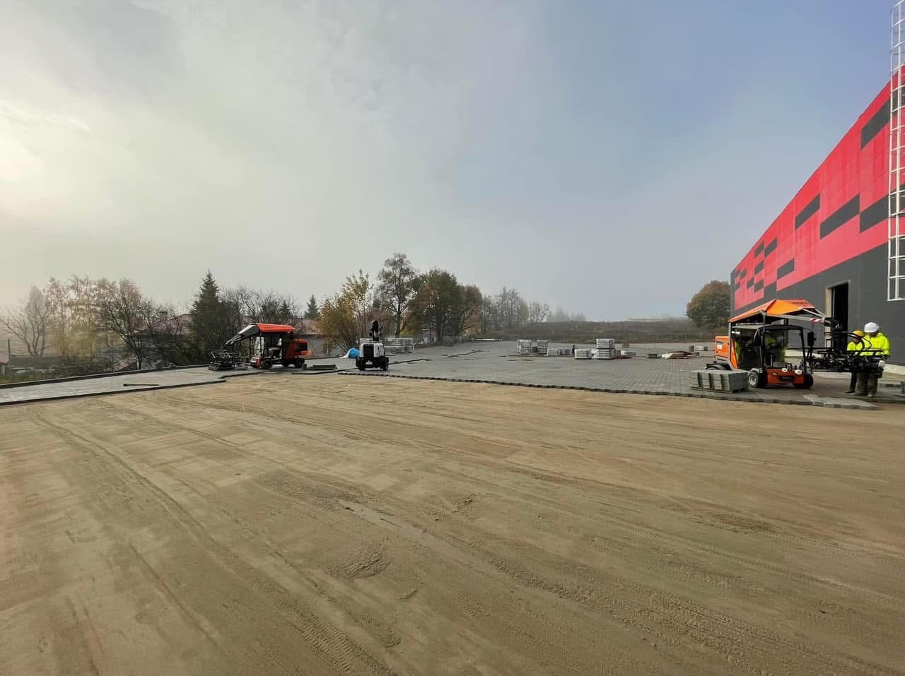 Rozległy plac z częściowo ułożoną szarą kostką brukową, maszyny do układania kostki i palety z kostką w tle, obok budynku z czerwono-czarną fasadą w pochmurny dzień.