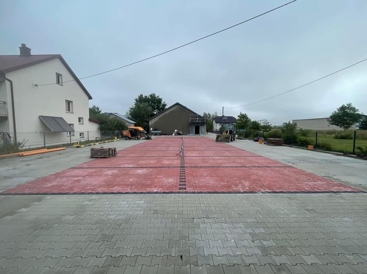 Rozległy plac z wyznaczonymi miejscami parkingowymi, częściowo wyłożony czerwoną kostką brukową, z widocznymi maszynami budowlanymi w tle, w pochmurny dzień.