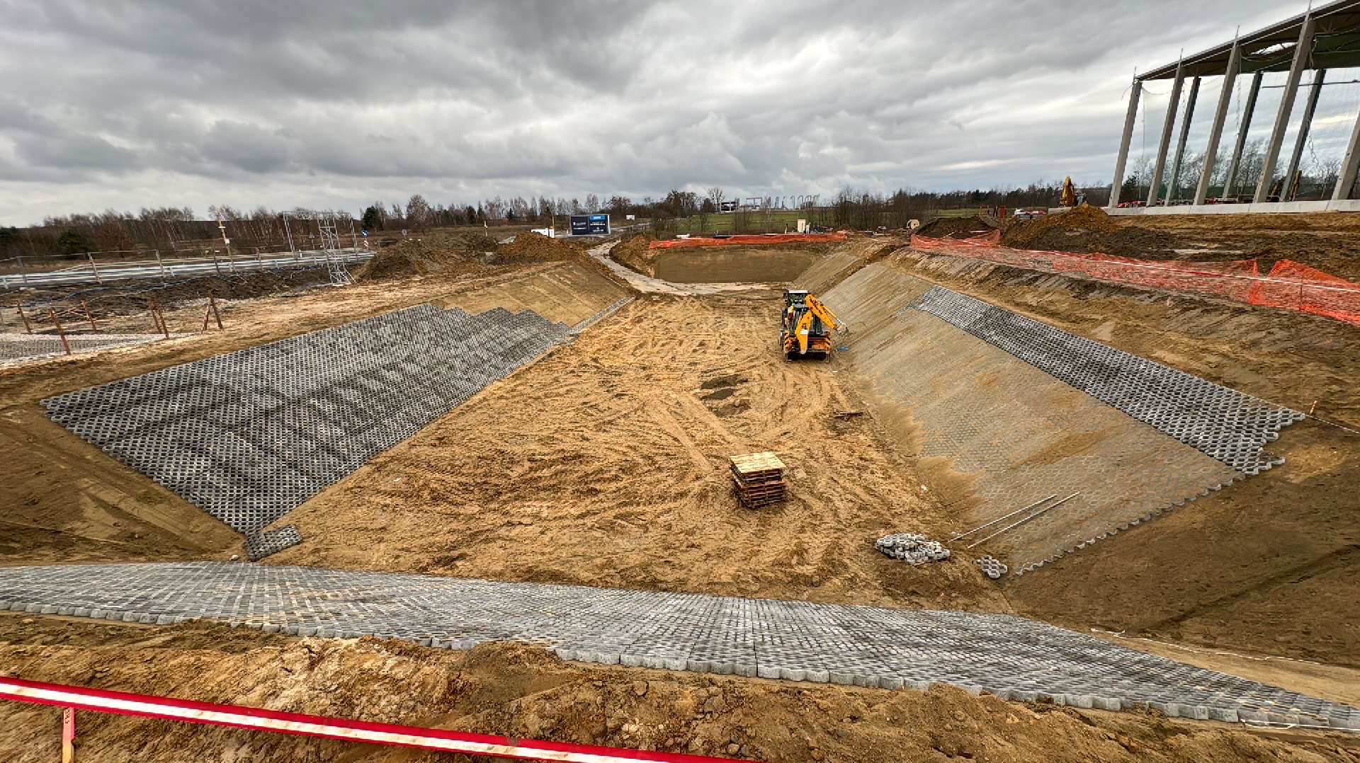 Szeroki wykop z częściowo ułożonymi ażurowymi płytami betonowymi na zboczach, żółta koparka w centralnej części wykopu, paleta z płytami i narzędzia obok, w tle budynek w trakcie budowy i pochmurne...