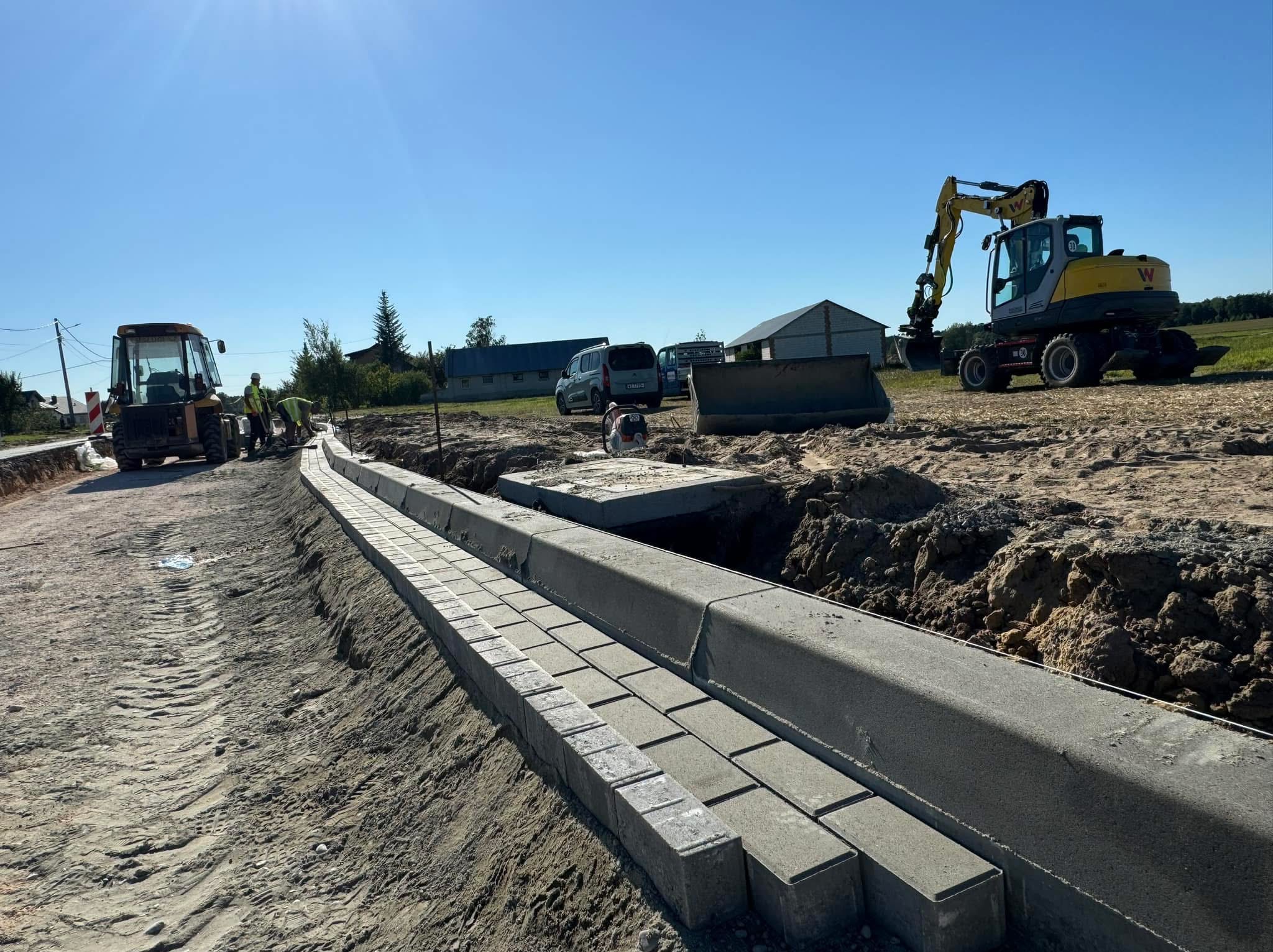 Świeżo ułożone szare kostki betonowe tworzą krawężnik wzdłuż drogi gruntowej, z widocznymi śladami opon po ciężkim sprzęcie, w tle żółta koparka i buldożer oraz pracownicy w kamizelkach...