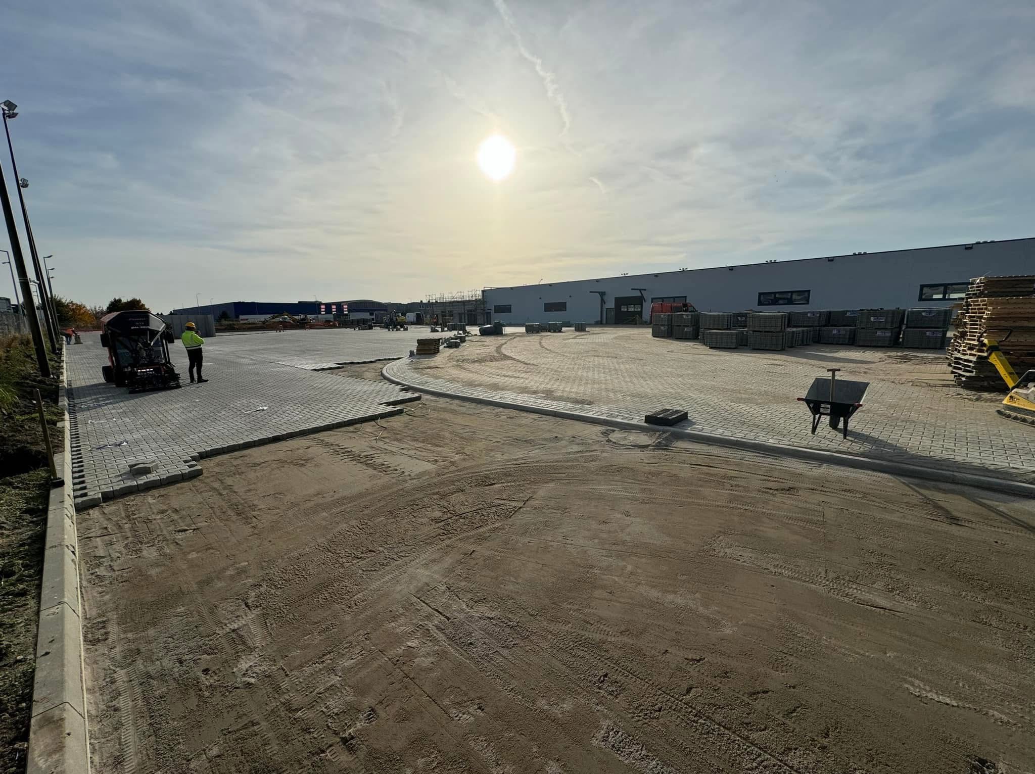 Układanie kostki brukowej na dużej powierzchni, widoczny pracownik z maszyną do układania kostki, palety z kostką oraz fragment już ułożonej nawierzchni. W tle budynek magazynowy w pochmurny dzień.