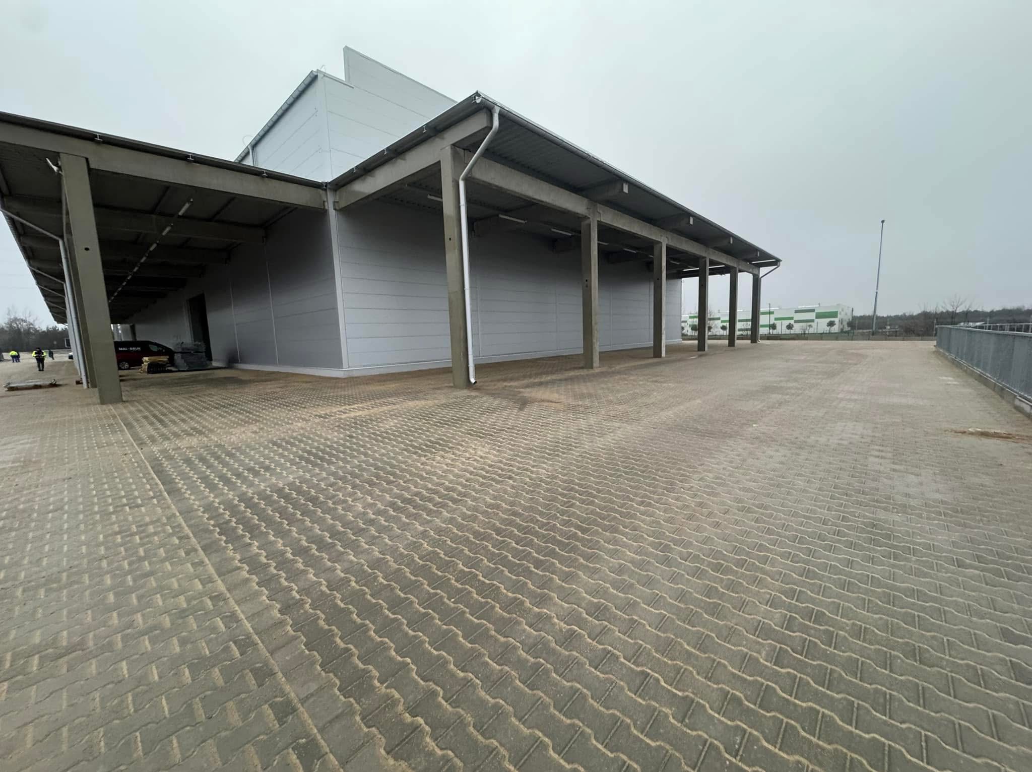 Szeroki kadr na dużą powierzchnię wyłożoną szarą kostką brukową przed magazynem z betonowymi słupami podtrzymującymi zadaszenie, widoczni pracownicy w oddali.
