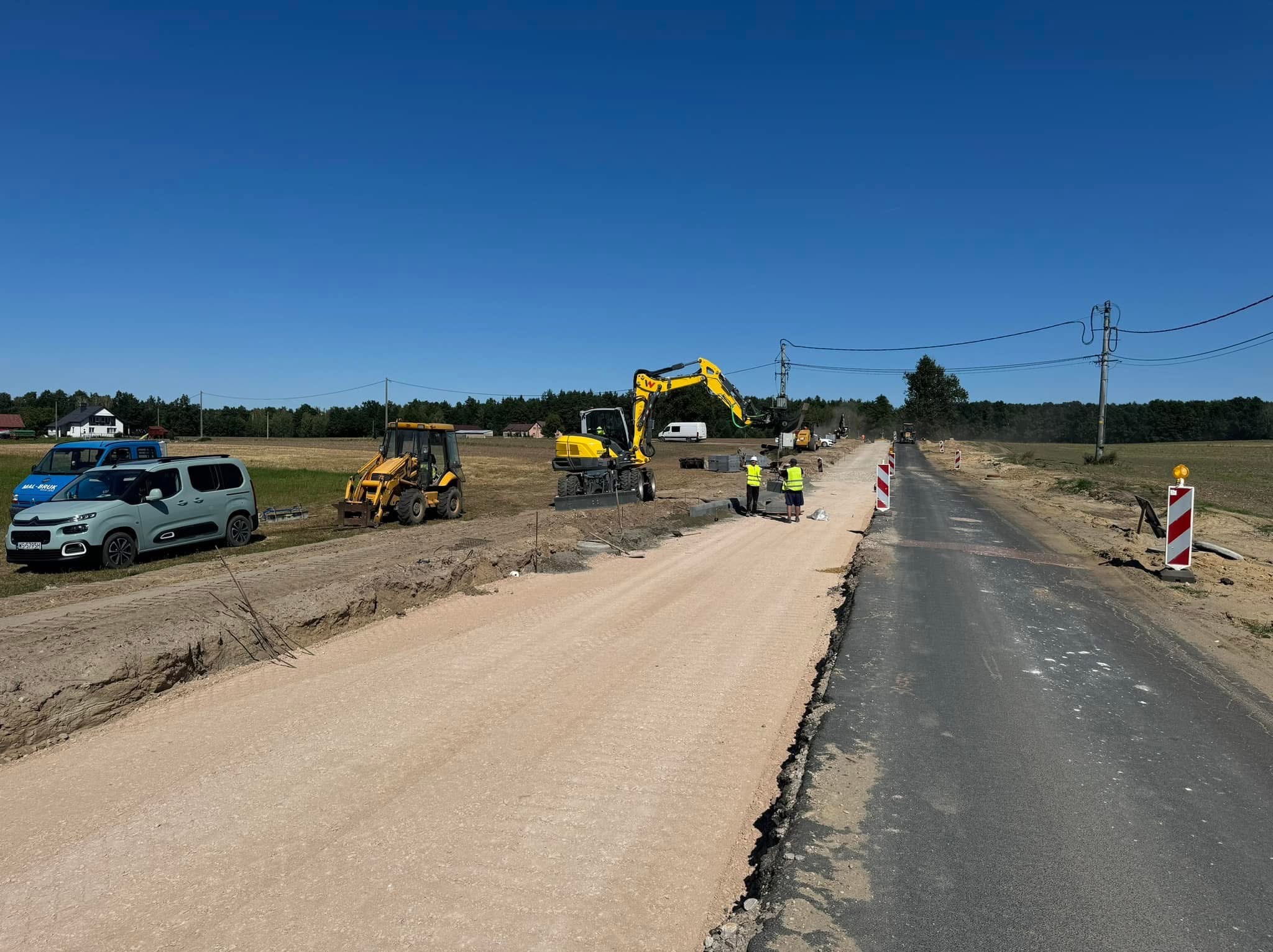 Żółty minikoparka i inne maszyny budowlane na placu budowy drogi z widocznym podkładem z kruszywa i fragmentem nawierzchni asfaltowej, w tle dom i błękitne auto.