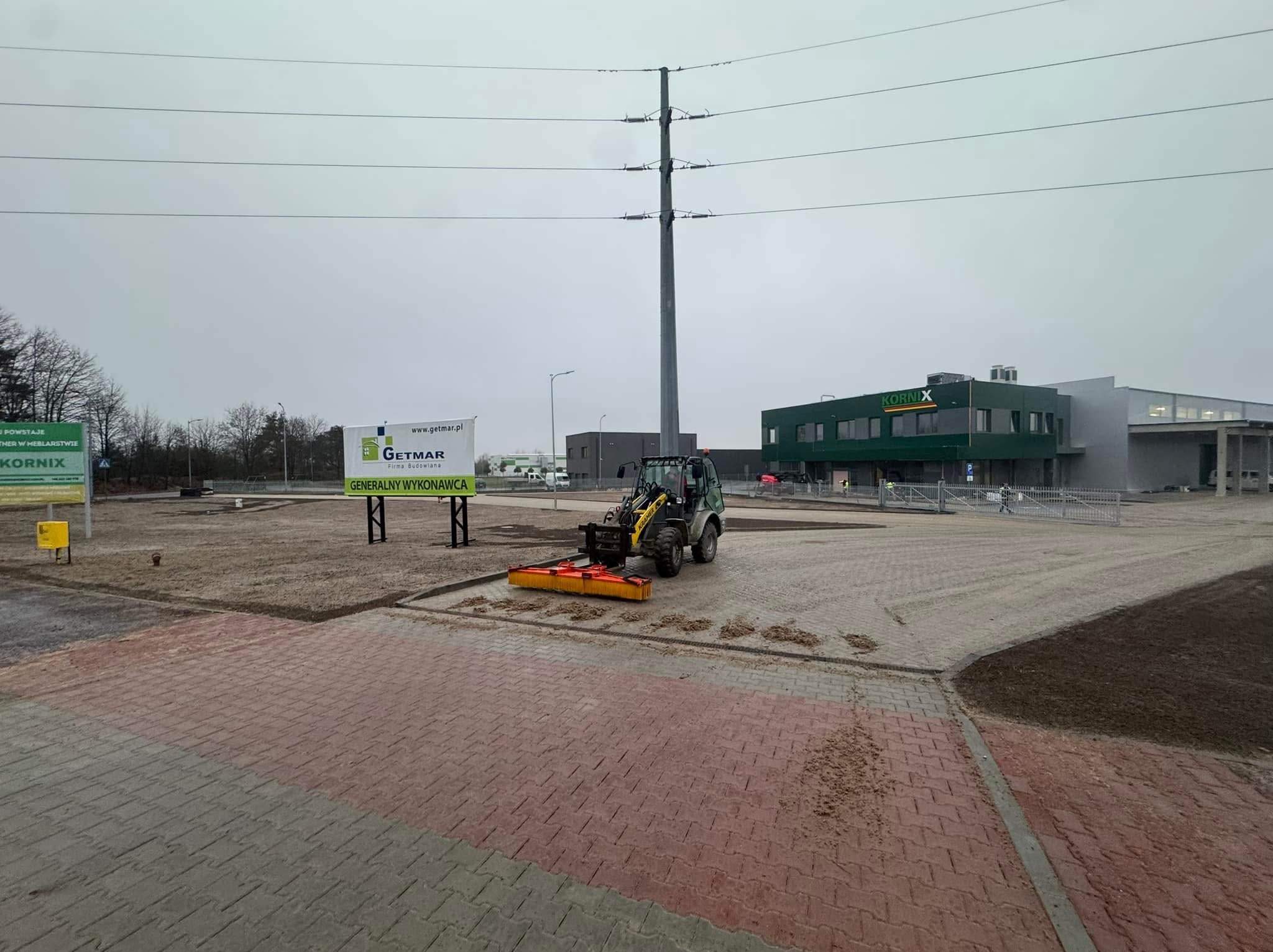 Kompaktowa zamiatarka na kostce brukowej, obok świeżo wyrównany teren pod budowę, w tle budynek firmy Kornix i słup wysokiego napięcia.
