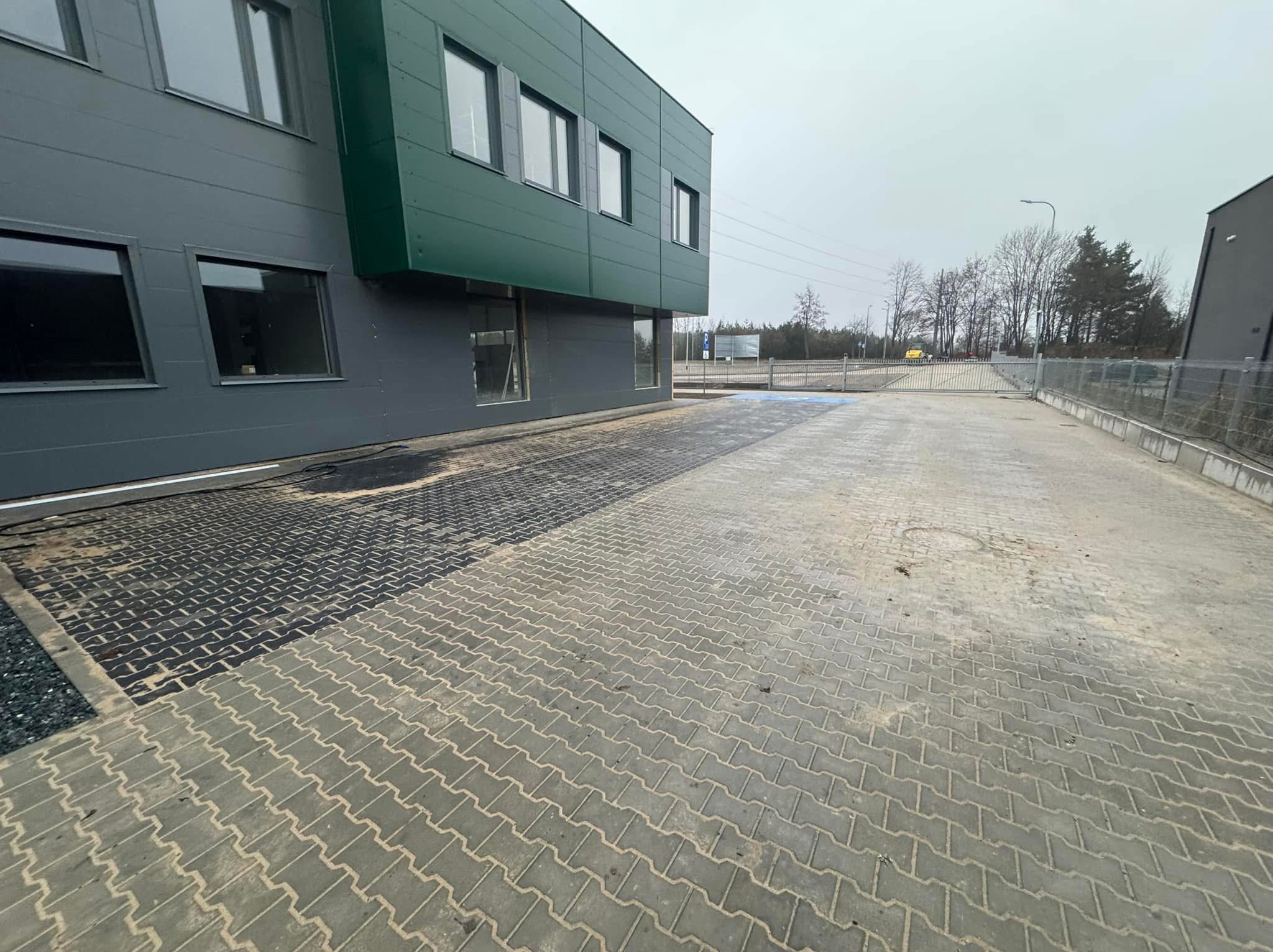 Nowo ułożona szara kostka brukowa przed nowoczesnym budynkiem biurowym z zielonymi akcentami, część nawierzchni jeszcze nieoczyszczona, widoczne ślady po pracach, w tle brama wjazdowa i drzewa.