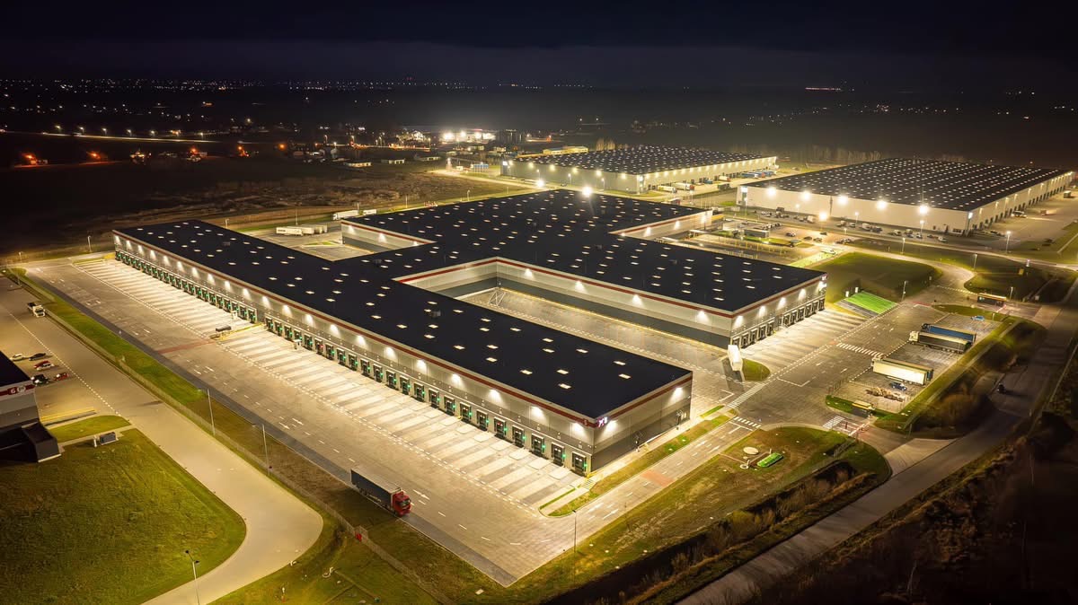 Nocny widok z lotu ptaka na kompleks magazynowy z rampami załadunkowymi, oświetlonymi parkingami i drogami dojazdowymi, w tle rozświetlone miasto.