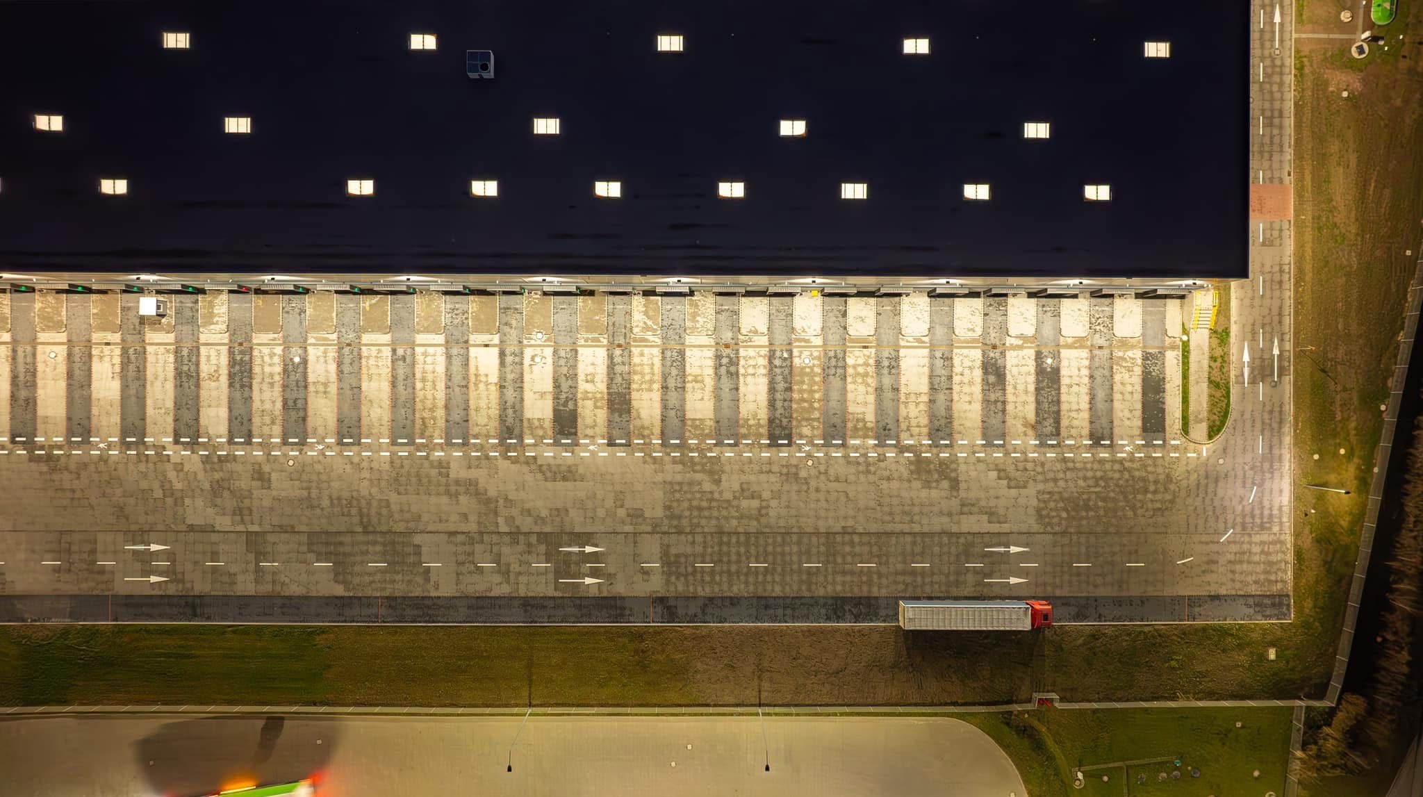 Widok z drona na plac rozładunkowy magazynu z zaparkowaną ciężarówką, widoczne doki rozładunkowe, oznakowanie poziome i oświetlenie nocne.