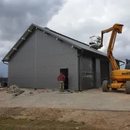 Szary budynek z płyt warstwowych z podnośnikiem koszowym i osobą w drzwiach; widoczne narzędzia i materiały budowlane na placu