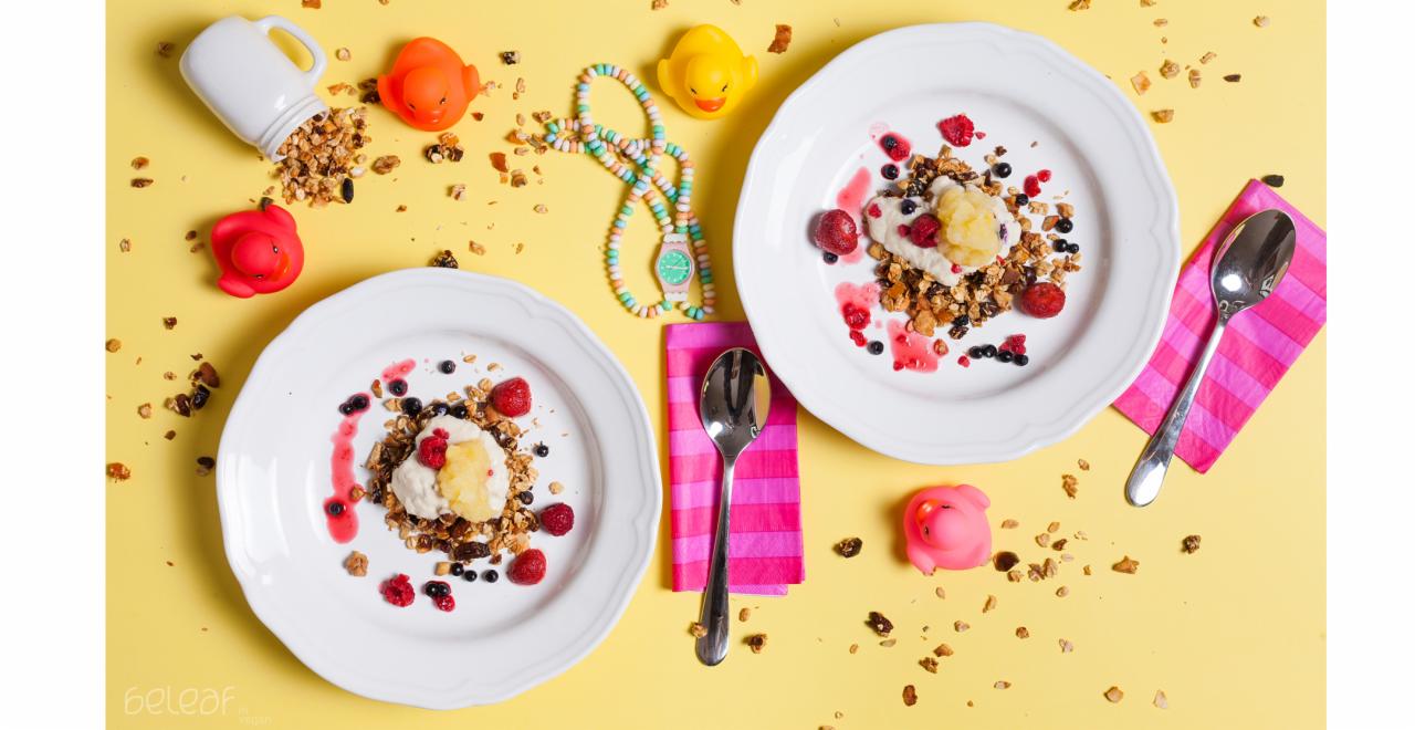 Dwa talerze z granolą, owocami (maliny, jagody, kawałki ananasa) i sosem na żółtym tle, otoczone rozsypaną granolą, gumowymi kaczkami, różowym prosiaczkiem, cukierkami i przewróconym kubkiem...