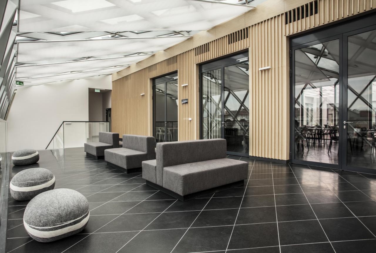 Nowoczesna poczekalnia w Luboniu z szarymi sofami modułowymi, pufami w kształcie kamieni i podłogą z czarnego kamienia z białymi fugami, oświetlona naturalnym światłem przez przeszklony sufit.