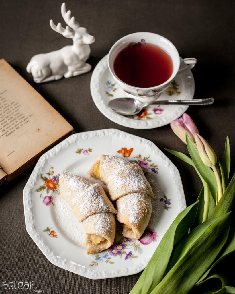 Dwa rogaliki z nadzieniem, posypane cukrem pudrem na talerzu z kwiatowym wzorem, obok filiżanka herbaty, biały ceramiczny renifer, tulipany i otwarta książka z widocznym tekstem.