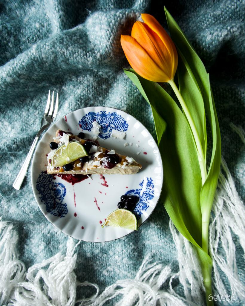 Kawałek tarty z owocami i limonką na talerzu z niebieskim wzorem, obok tulipan i srebrny widelec, całość na tle turkusowego koca z frędzlami.