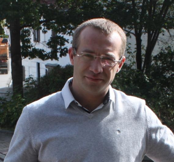 Portret mężczyzny w okularach i szarym swetrze na tle drzew i budynku, z lekkim uśmiechem.