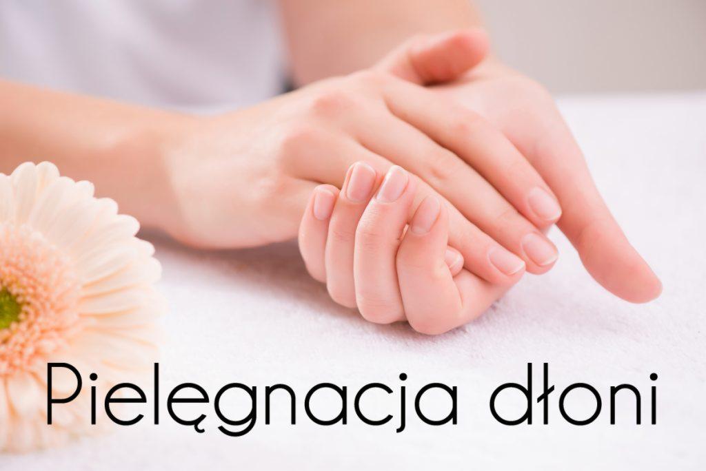 Dłonie kobiety z naturalnym manicure, sfotografowane na białym ręczniku obok jasnej gerbery. Tekst 'Pielęgnacja dłoni' na pierwszym planie.