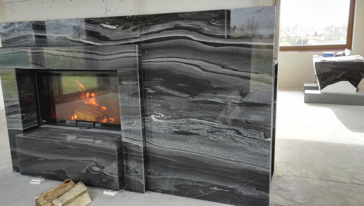 Nowoczesna obudowa paleniska z czarnego kamienia z wyraźnym wzorem żyłkowania, płomienie wewnątrz, drewno obok, surowy stan wykończenia wnętrza.