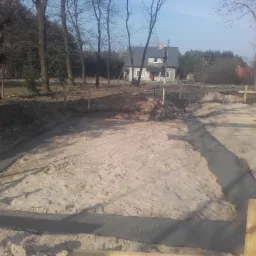 Przygotowanie terenu pod budowę domu w Chorchosach: wytyczone fundamenty z wylanym betonem, widok na działkę z piaskiem i pobliskie drzewa.