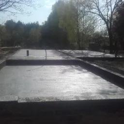 Świeżo wylana, gładka płyta fundamentowa pod dom jednorodzinny, z widocznymi pionowymi prętami zbrojeniowymi i wystającą rurą instalacyjną, otoczona ziemią i drzewami w tle.