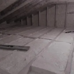 Izolacja poddasza wełną mineralną między drewnianymi belkami, widoczna poziomica i narzędzia, surowe wykończenie ścian z płyt OSB.