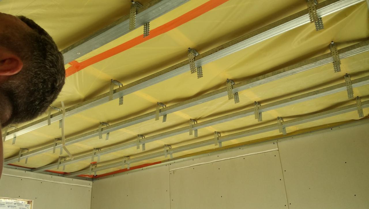 Montaż sufitu podwieszanego: widoczne profile metalowe, izolacja z wełny mineralnej i fragment głowy pracownika, ściany z płyt kartonowo-gipsowych.