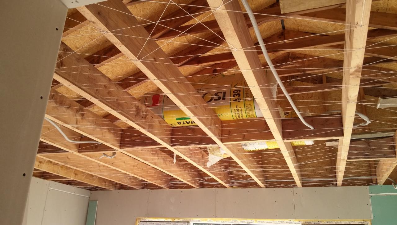 Montaż sufitu podwieszanego: drewniana konstrukcja z widoczną izolacją w rolce, przewody elektryczne i naciągnięte sznurki poziomujące, częściowo zamontowane płyty gipsowo-kartonowe.