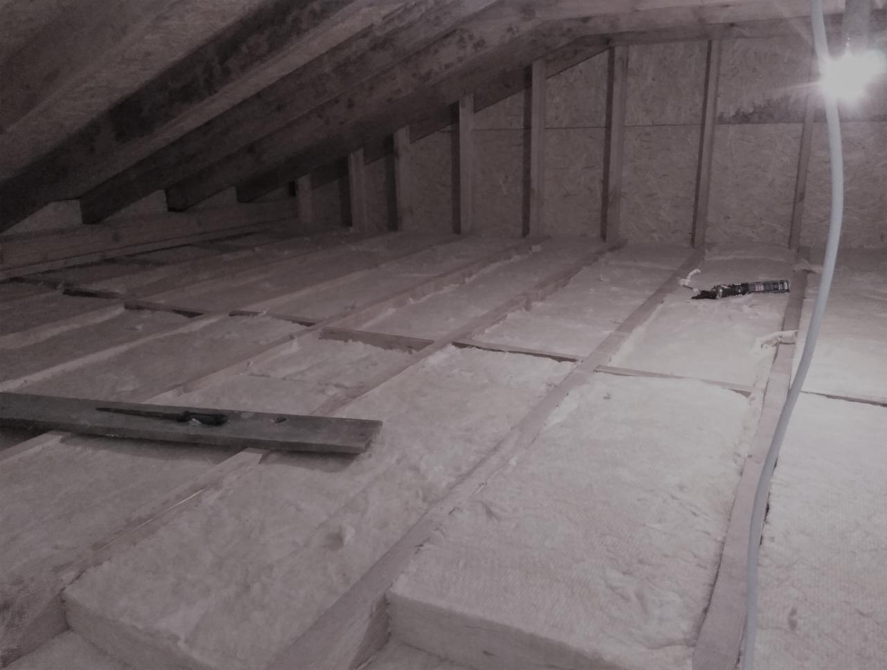 Izolacja poddasza wełną mineralną między drewnianymi belkami, widoczna poziomica i narzędzia, surowe wykończenie ścian z płyt OSB.