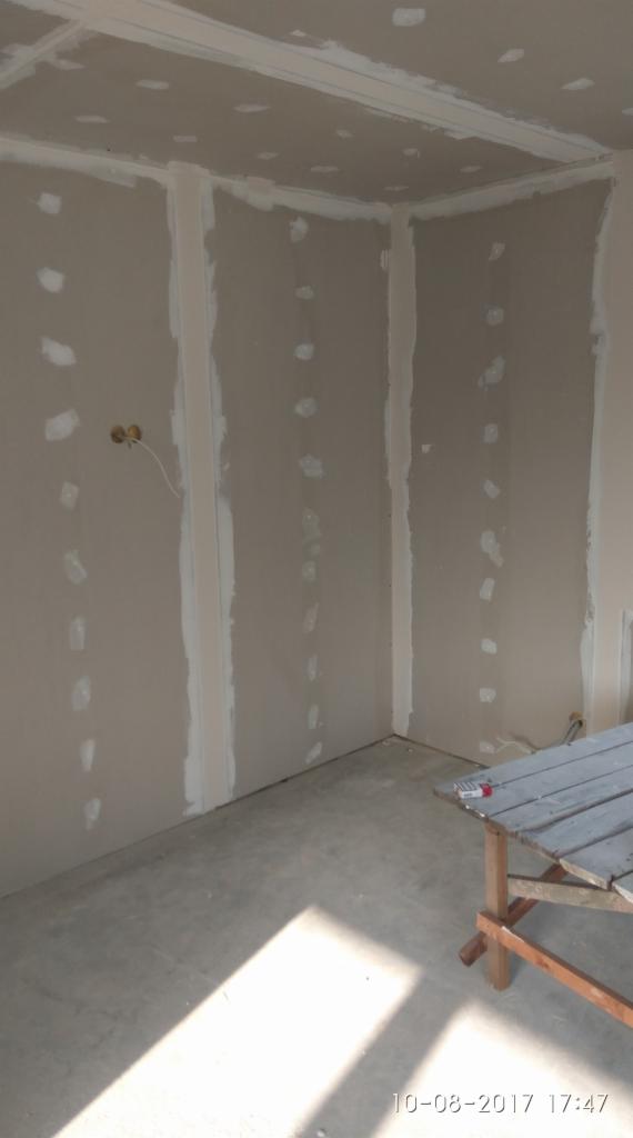 Wnętrze w trakcie prac wykończeniowych: ściany z płyt gipsowo-kartonowych, z widocznymi miejscami po wkrętach, wstępnie zaszpachlowane, przygotowane do dalszej obróbki, na podłodze widoczny cień,...