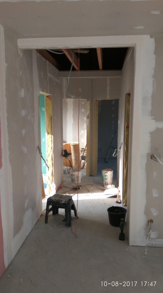 Wnętrze w trakcie remontu w Garwolinie: surowe ściany przygotowane do malowania, widoczne instalacje elektryczne, podłoga wylewka, czarny stołek i wiadro na podłodze, w tle materiały izolacyjne.