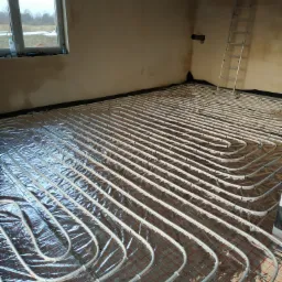 Białe rury ogrzewania podłogowego ułożone na folii odblaskowej z czerwoną siatką, widok na surowe ściany i okno z widokiem na zimowy krajobraz, drabina oparta o ścianę w tle.