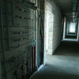 Szereg rur instalacyjnych z zaworami zamontowanych na ścianie z bloczków betonowych w surowym stanie, widok na długi korytarz z instalacjami pod stropem.