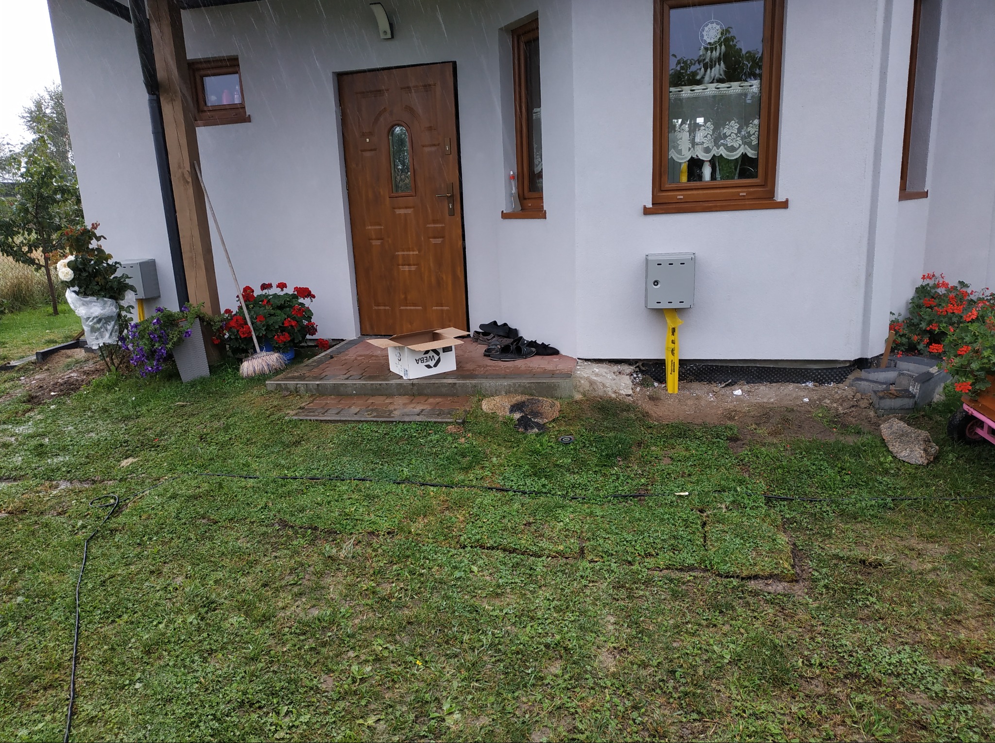 Prace wykończeniowe w Koszalinie: świeżo otynkowany dom z brązowymi oknami i drzwiami wejściowymi, widoczne kwiaty w donicach i skrzynka instalacyjna gazu na elewacji. Na trawniku widoczny kabel...