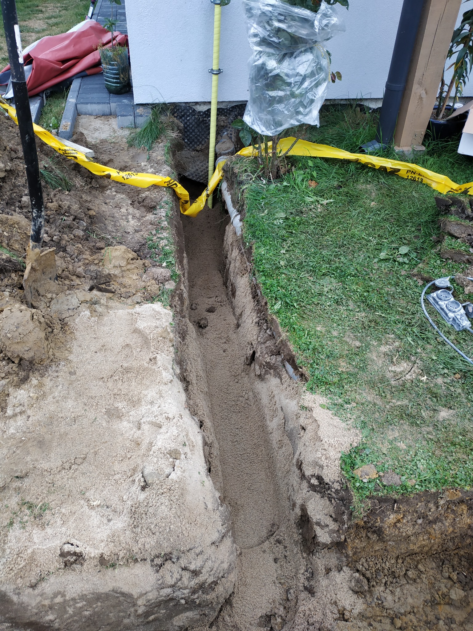 Wykop liniowy w ziemi, zabezpieczony żółtą taśmą ostrzegawczą, prawdopodobnie pod instalację gazową lub wodną, z widocznymi elementami instalacji i fragmentem budynku w tle.