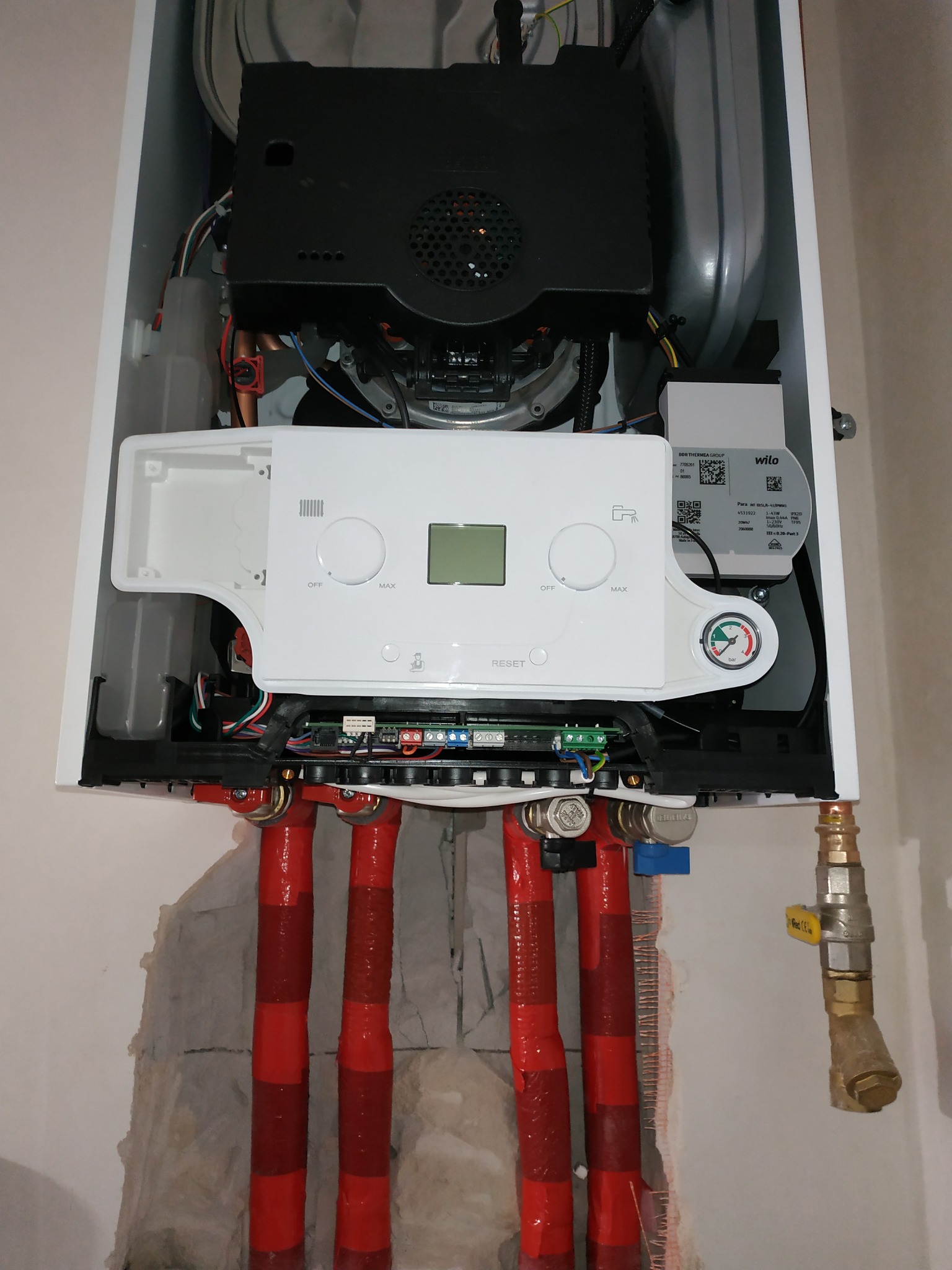 Otwarty piec gazowy z widocznymi czerwonymi rurami instalacji grzewczej i elementami sterującymi, zamontowany na ścianie z widocznymi śladami prac instalacyjnych.