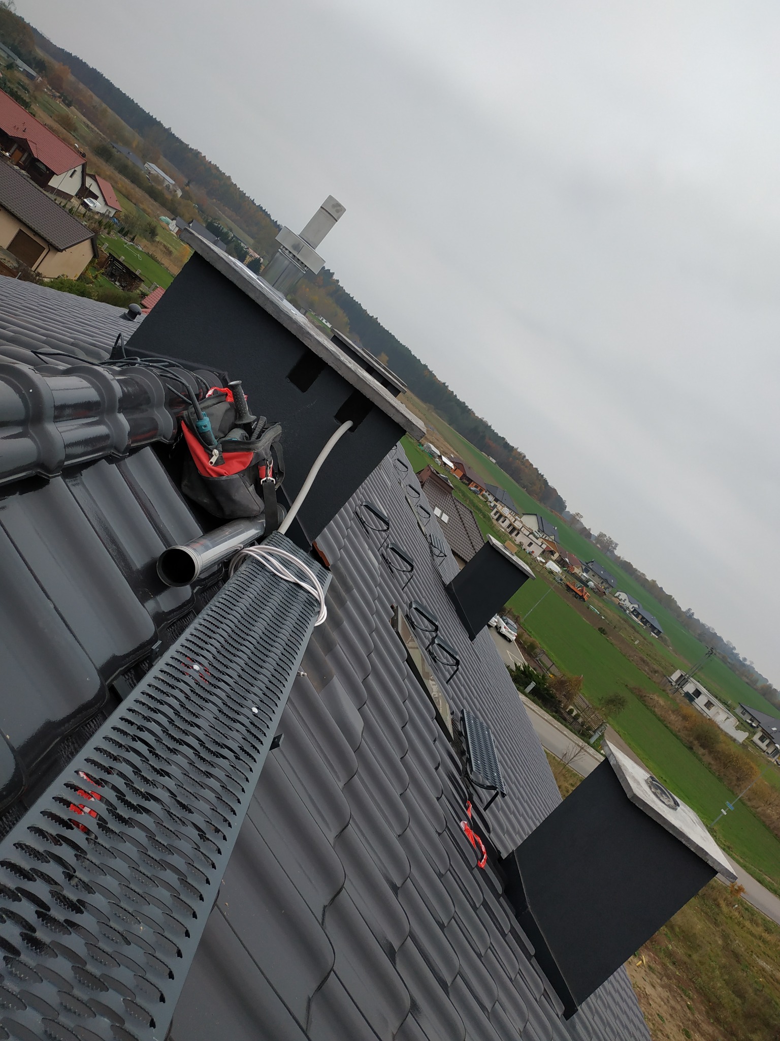 Ciemna dachówka ceramiczna z zamontowanym kominem i ławą kominiarską, widoczny przewód wentylacyjny i torba narzędziowa w kolorze czerwonym, w tle zabudowa jednorodzinna i pochmurne niebo.
