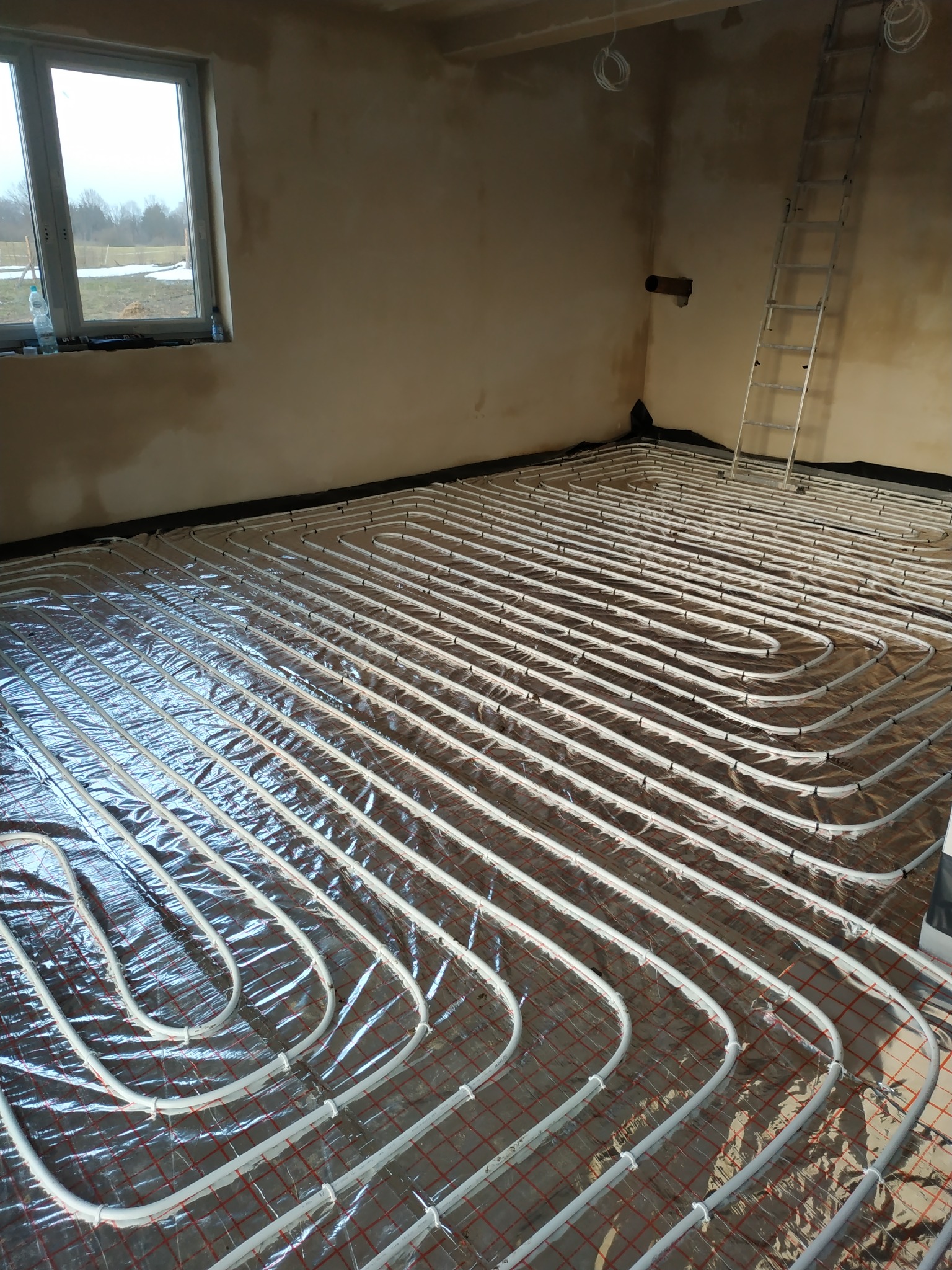 Białe rury ogrzewania podłogowego ułożone na folii odblaskowej z czerwoną siatką, widok na surowe ściany i okno z widokiem na zimowy krajobraz, drabina oparta o ścianę w tle.
