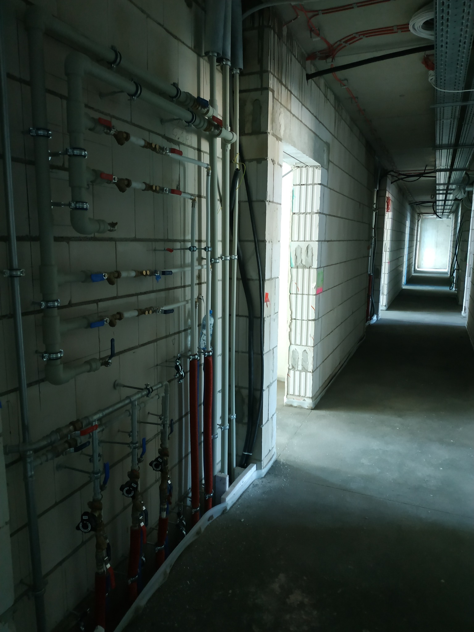 Szereg rur instalacyjnych z zaworami zamontowanych na ścianie z bloczków betonowych w surowym stanie, widok na długi korytarz z instalacjami pod stropem.