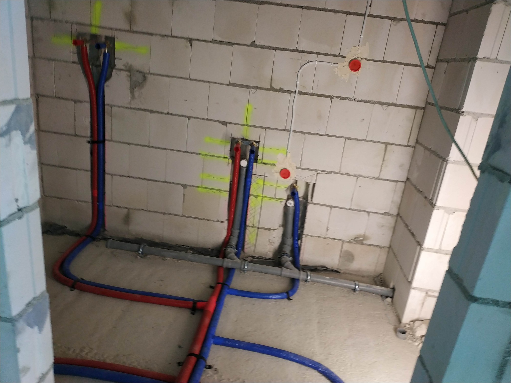 Surowe ściany z betonu komórkowego z zaznaczonymi punktami instalacji wodnej (rury czerwone i niebieskie) i elektrycznej (białe kable), widoczne szare rury kanalizacyjne.