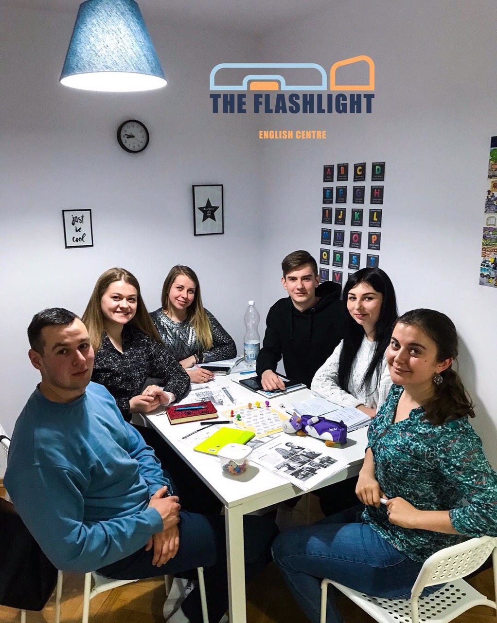 Grupa uczniów i nauczyciel w klasie we Wrocławiu, siedząca przy białym stole z materiałami edukacyjnymi, logo 'The Flashlight English Centre' na ścianie.