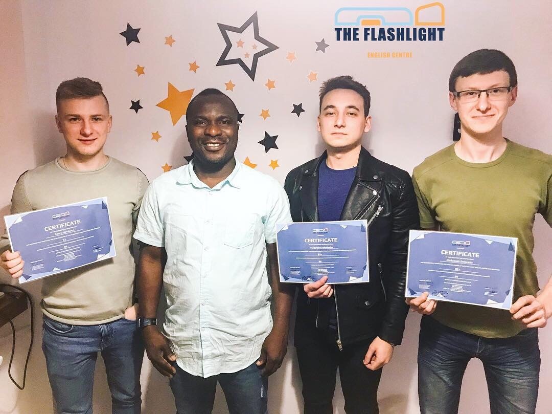 Grupa czterech osób, w tym jeden czarnoskóry mężczyzna, prezentuje certyfikaty ukończenia kursu językowego 'The Flashlight English Centre' na tle ściany z dekoracyjnymi gwiazdami.