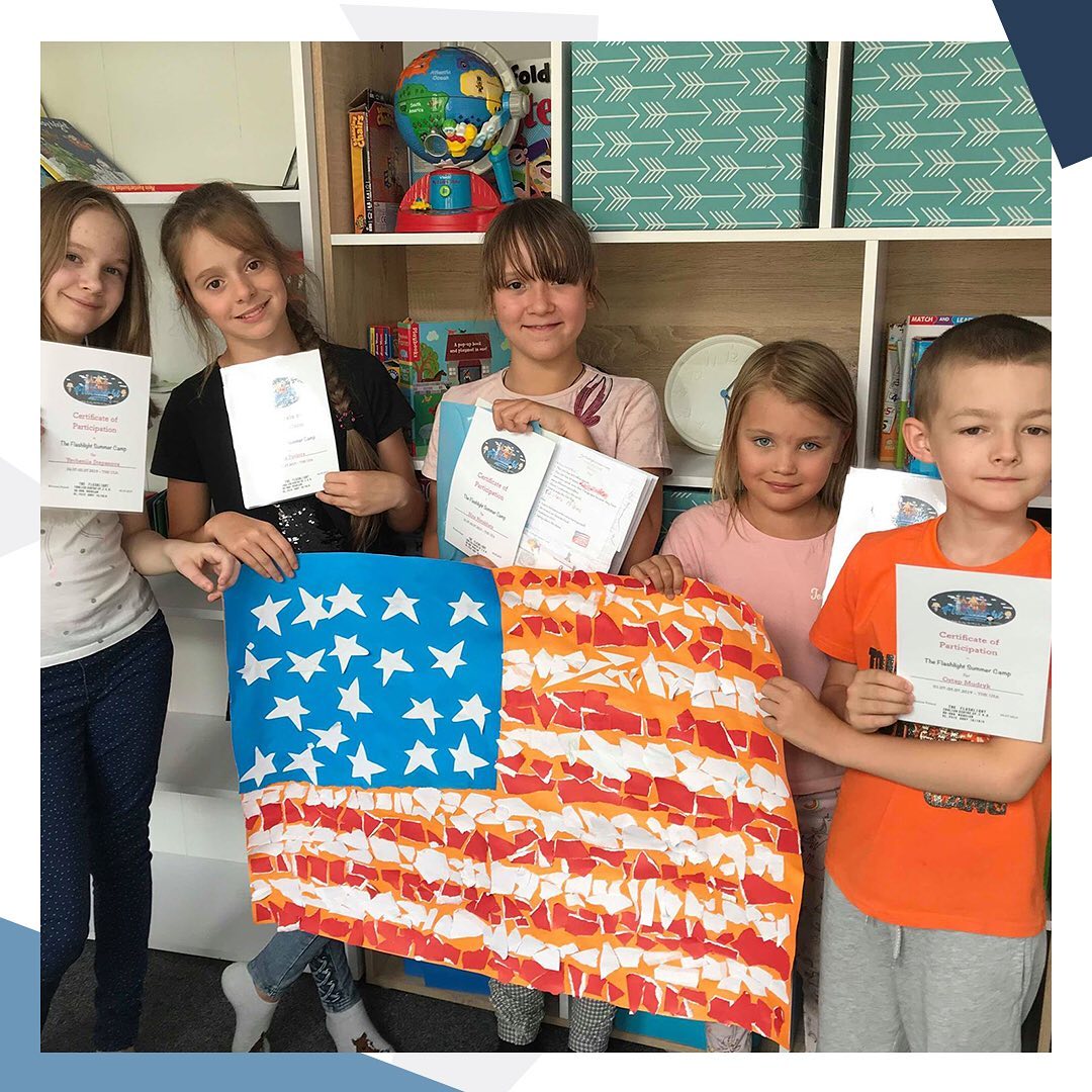 Grupa dzieci trzymająca dyplomy i flagę USA wykonaną z papierowych kawałków na tle półki z książkami i globusem.