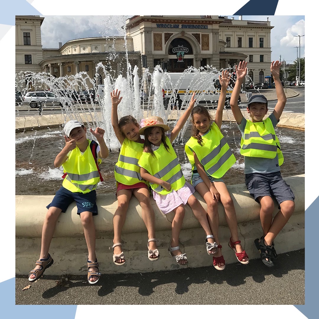 Pięcioro uśmiechniętych dzieci w odblaskowych kamizelkach siedzi na murku fontanny przed budynkiem Wrocław Świebodzki, machając rękami.