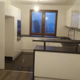 Biała kuchnia z czarnym blatem i oknem z drewnianą ramą w trakcie montażu, widoczne niedociągnięcia wykończeniowe.