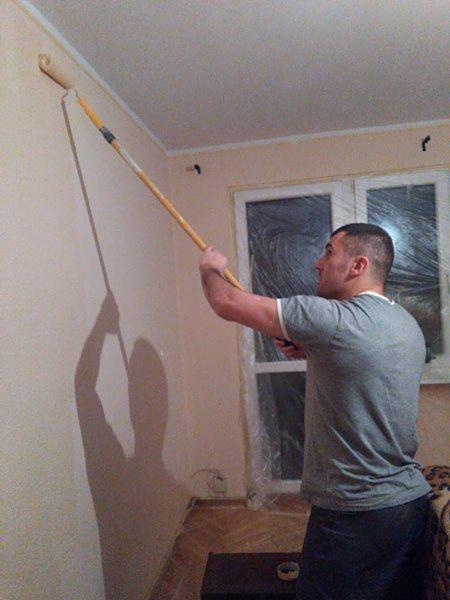 Mężczyzna maluje ścianę wałkiem na długim kiju, widoczny cień malarza na ścianie, okno zabezpieczone folią malarską.