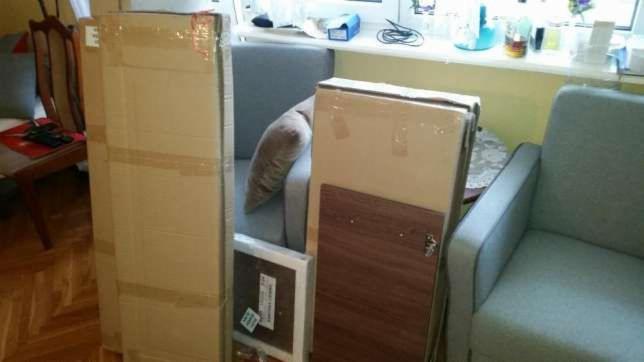 Paczki meblowe w kartonach, przygotowane do montażu w salonie, widoczne elementy drewniane i okucia.