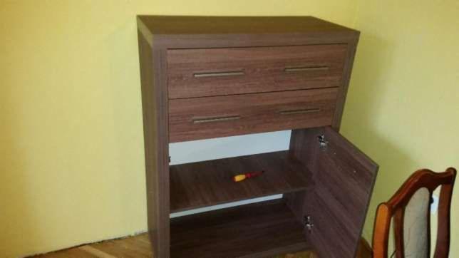 Brązowa komoda z szufladami i otwartymi drzwiczkami, na półce wewnątrz leży czerwono-żółty śrubokręt, obok stoi krzesło z drewnianym oparciem.