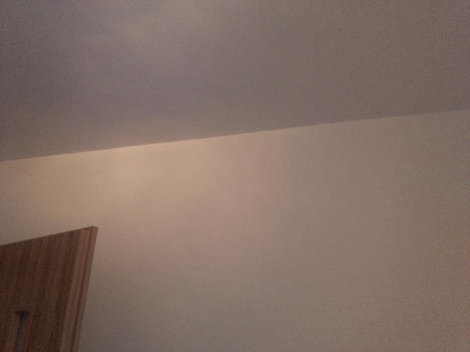 Pomalowana ściana w odcieniach beżu z fragmentem drewnianej ościeżnicy drzwiowej w lewym dolnym rogu.