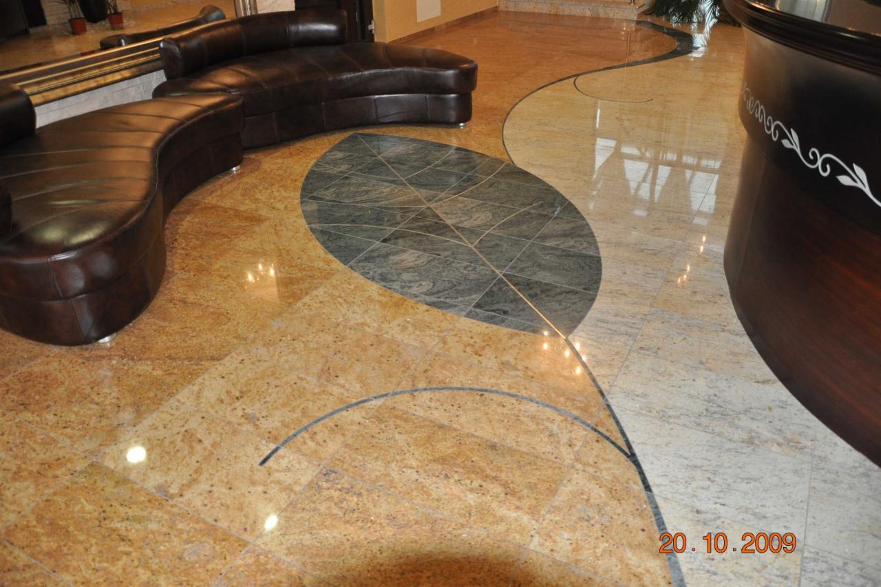 Dekoracyjna posadzka kamienna z motywem liścia wykonana z płytek w odcieniach szarości, otoczona przez złote płytki z widocznym fragmentem sofy ze skóry.