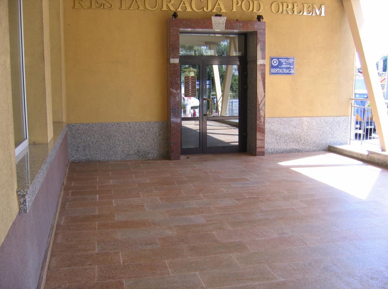 Wyłożona kamiennymi płytami posadzka przed wejściem do restauracji 'Pod Orłem', z widoczną granitową ramą drzwi wejściowych i fragmentem żółtej elewacji z szarym cokołem.