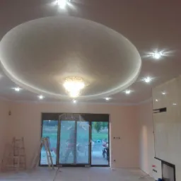 Owalny, podwieszany sufit z centralnym żyrandolem i punktowym oświetleniem LED, widoczny w trakcie remontu pomieszczenia z dwoma drabinami opartymi o okno tarasowe.