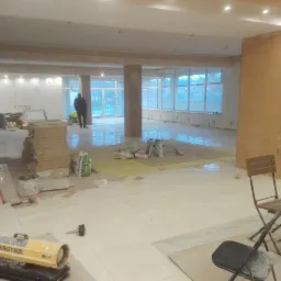 Rozległe wnętrze w trakcie prac remontowych, z widocznymi narzędziami, materiałami budowlanymi, oraz pracownikami w tle. Na pierwszym planie nagrzewnica MASTER i stos płytek podłogowych, co...