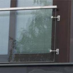 Zig zak elementy nierdzewne - Szklana balustrada balkonu z metalowymi mocowaniami, widok z zewnątrz, na tle szarej elewacji budynku z kamienia łupanego.